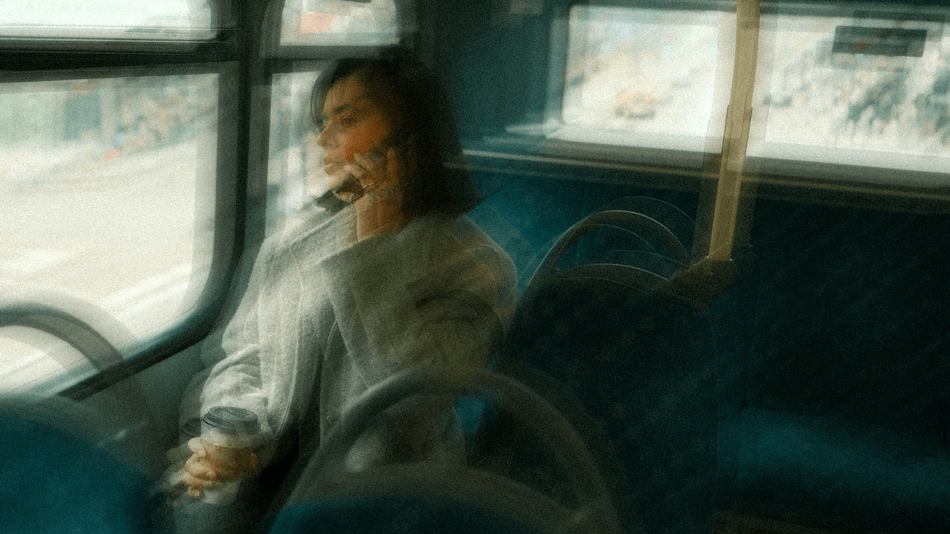 A woman in a coat holding a coffee cup, speaking on the phone while seated on a moving bus.