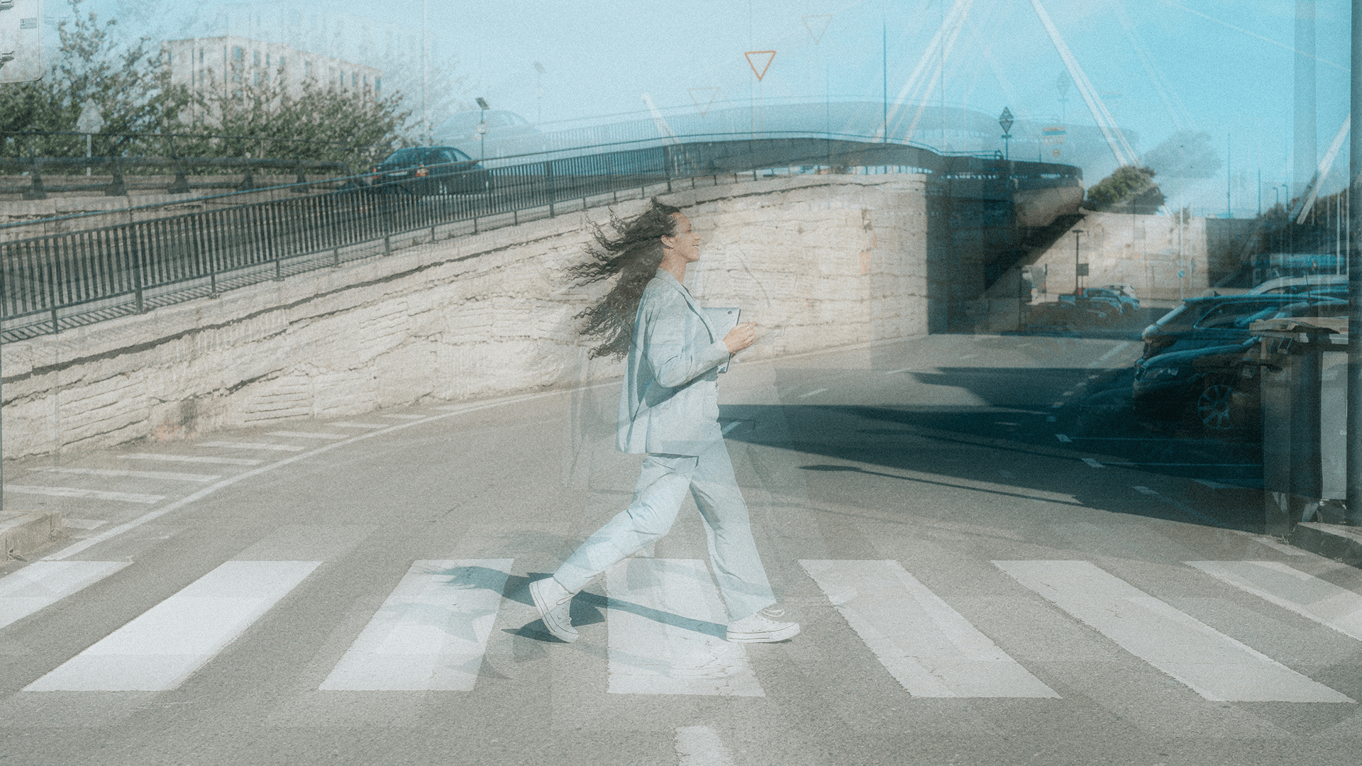 A woman in a light blue suit walking confidently across a city crosswalk, with motion blur emphasizing movement.